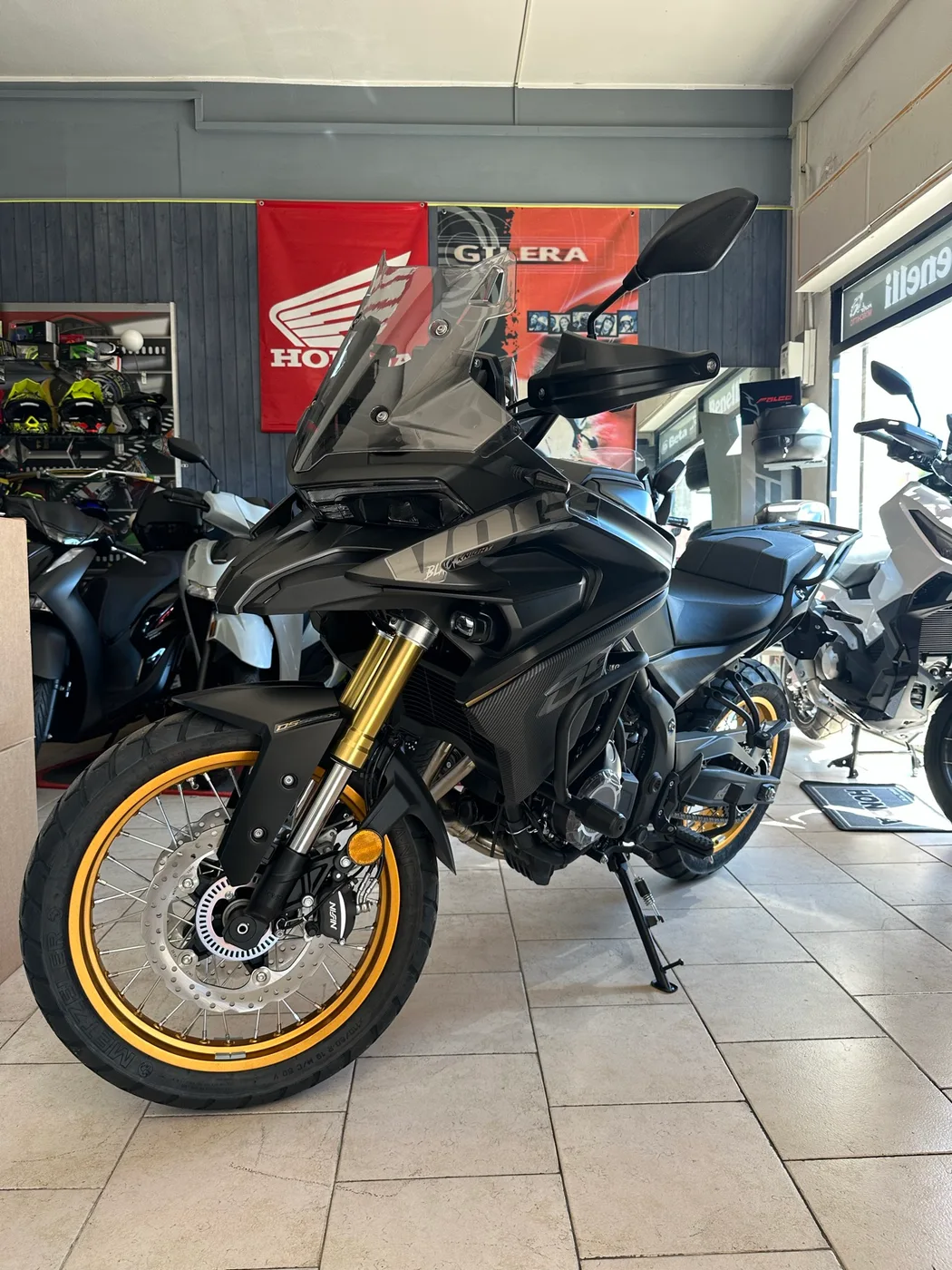 Moto adventure scura con cerchi dorati, fotografata nel negozio.